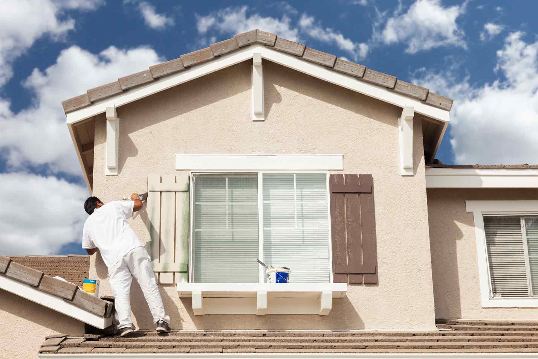 An exterior painter applying fresh paint to window shutters on a house